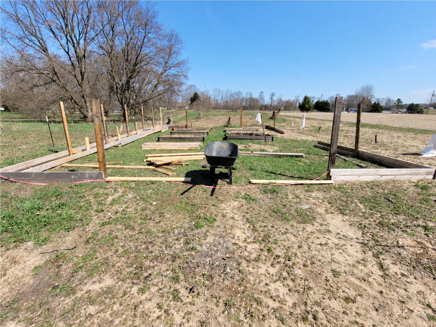 Wide view of the garden perimeter during construction, with a wheelbarrow, stacked lumber, and island beds.