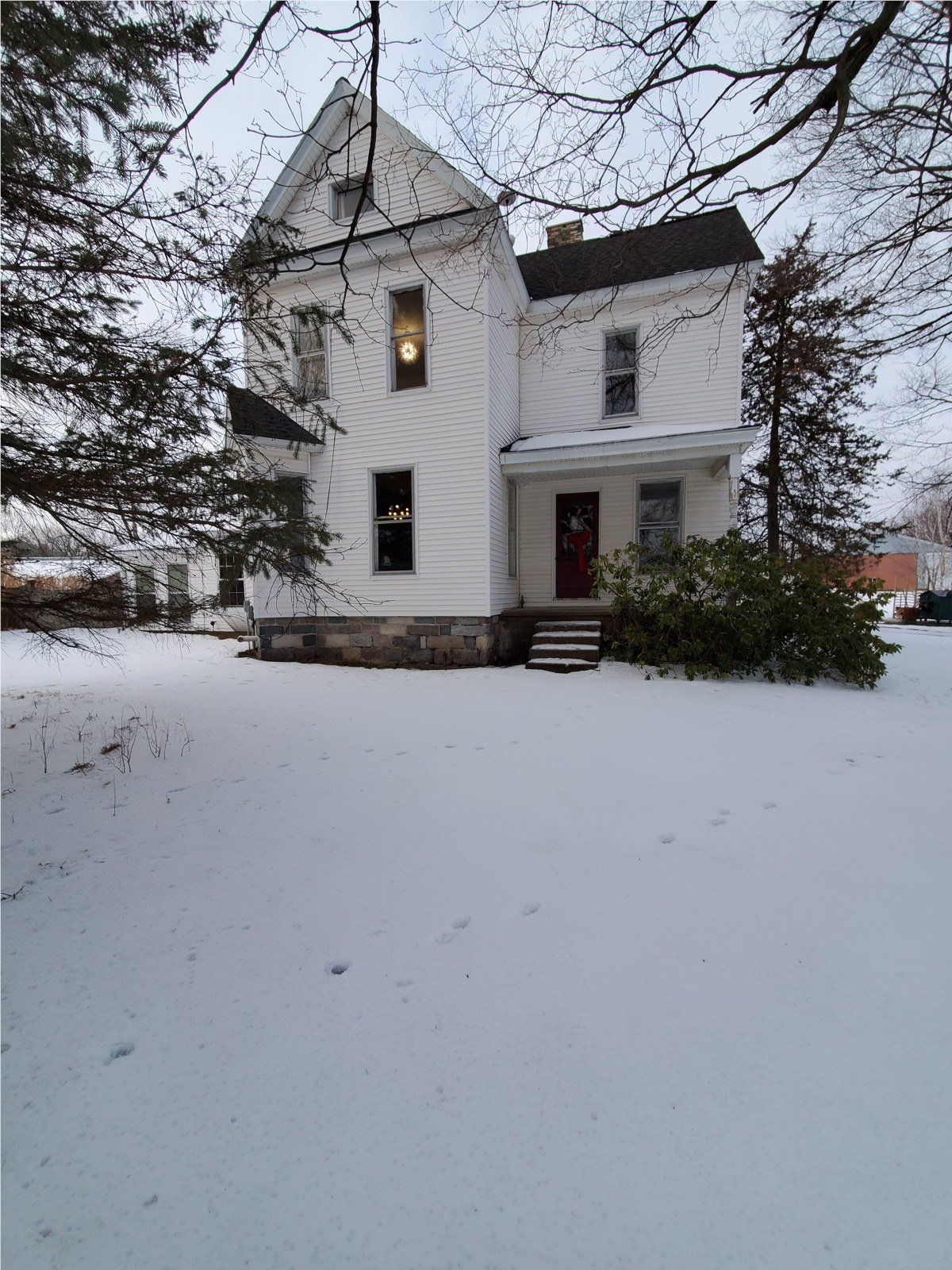 The white farmhouse in snow, with its red front door and a light glowing upstairs.